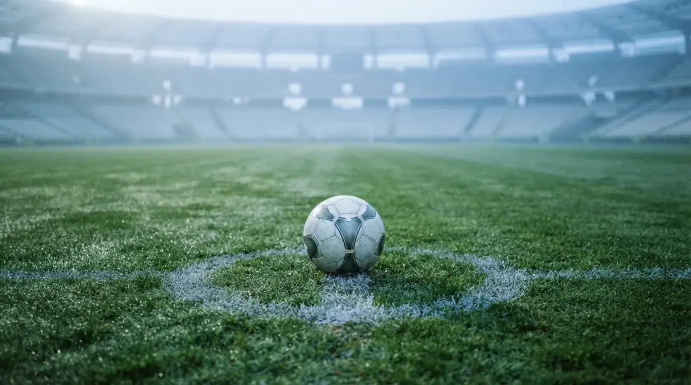 Ein einzelner Fussball liegt auf dem Anstosspunkt eines leeren Rasenplatzes am frühen Morgen, Tau glitzert auf dem Gras, die Tribüne liegt im Dunst.