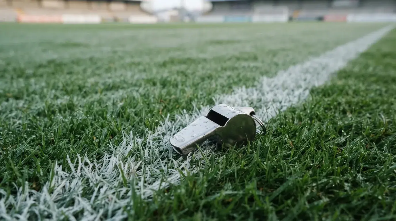 Eine Trillerpfeife liegt auf dem Rasen neben der Mittellinie, Tau hängt an den Halmen, im Hintergrund ein leerer Spielfeldrand.