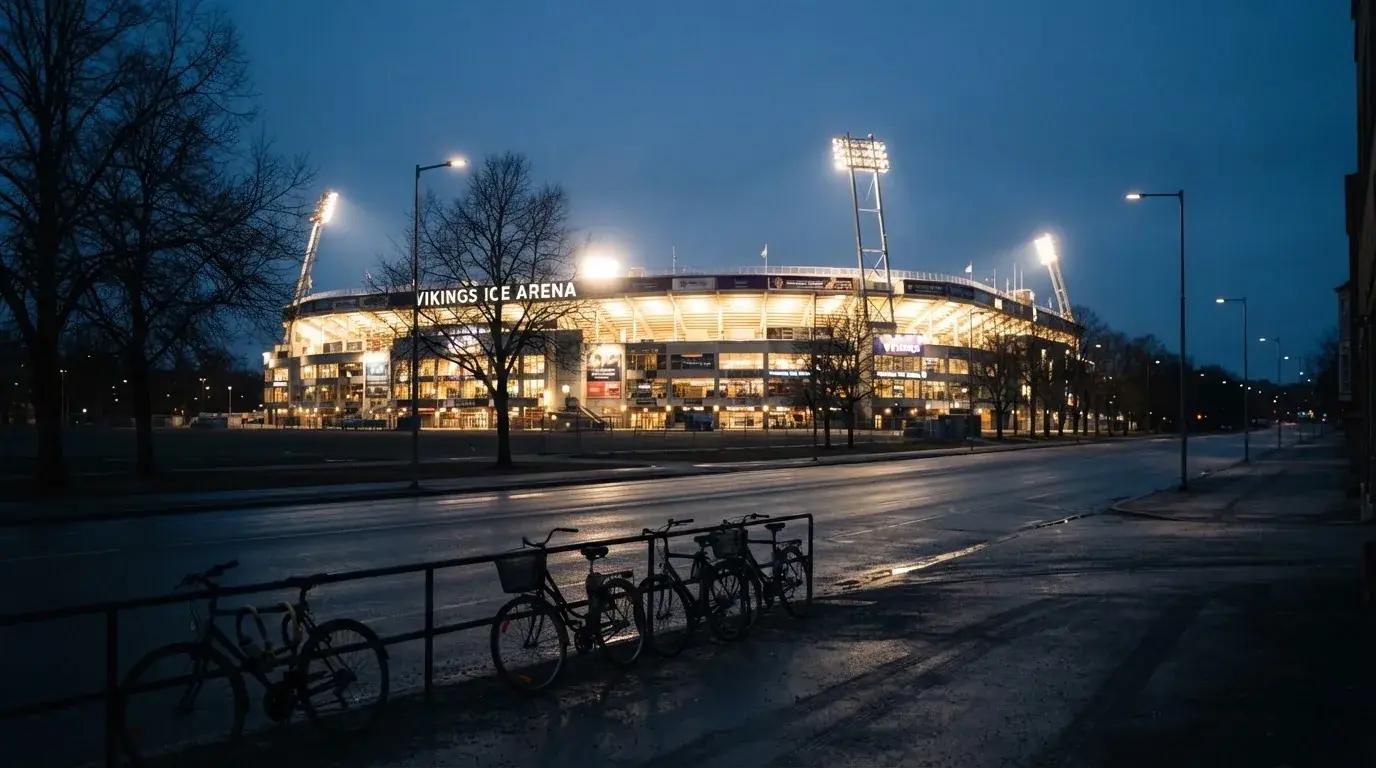 Ein grosses Eisstadion aus der Ferne gesehen, hell erleuchtet von Flutlicht in der Abenddämmerung, davor eine leere Strasse.