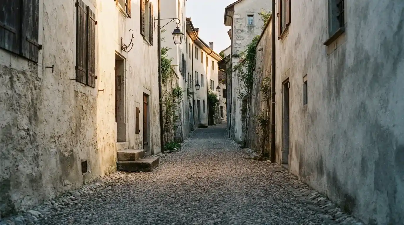 Ein schmaler Kiesweg führt zwischen zwei hellen Mauern durch eine Schweizer Altstadt, die Laternen sind noch nicht angezündet.