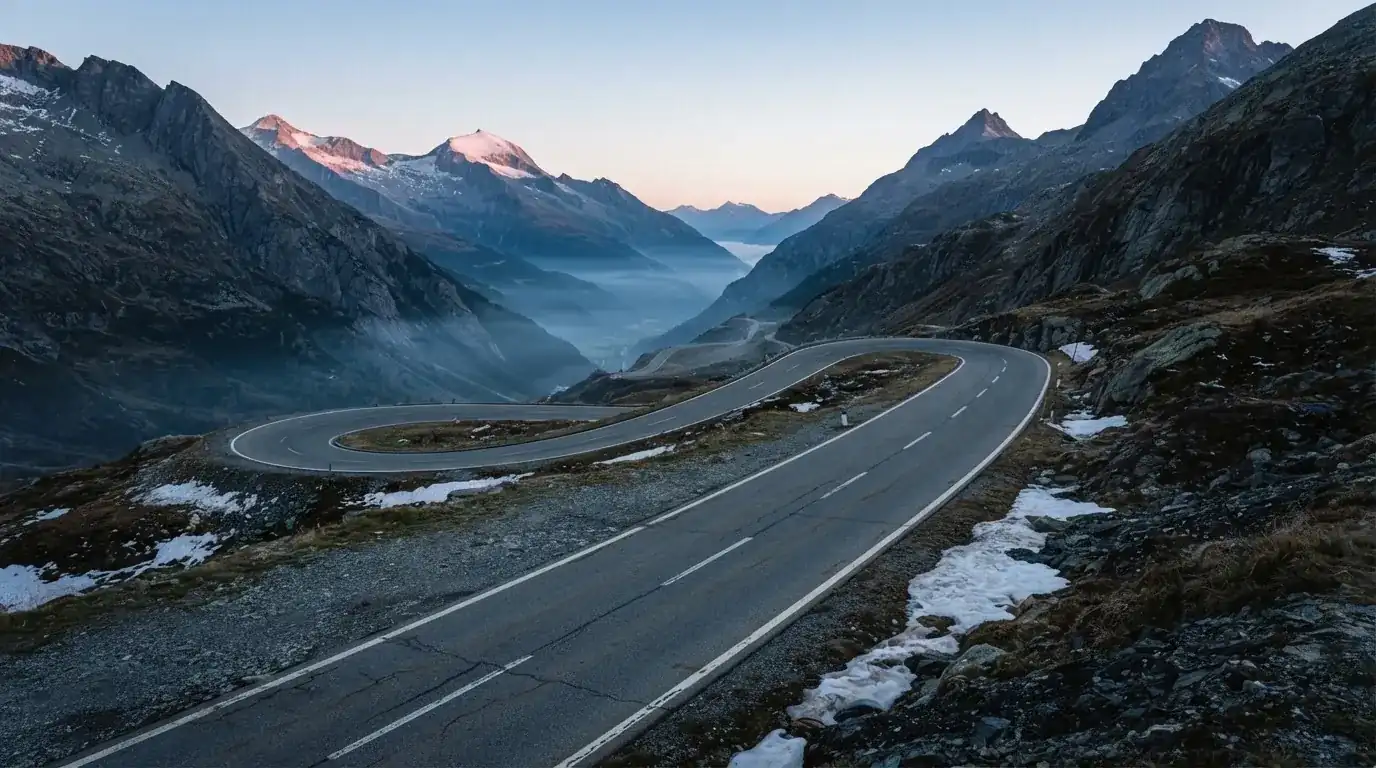 Ein Bergpass in den Schweizer Alpen mit leerer Strasse und Schneeresten am Rand im ersten Frühlicht.