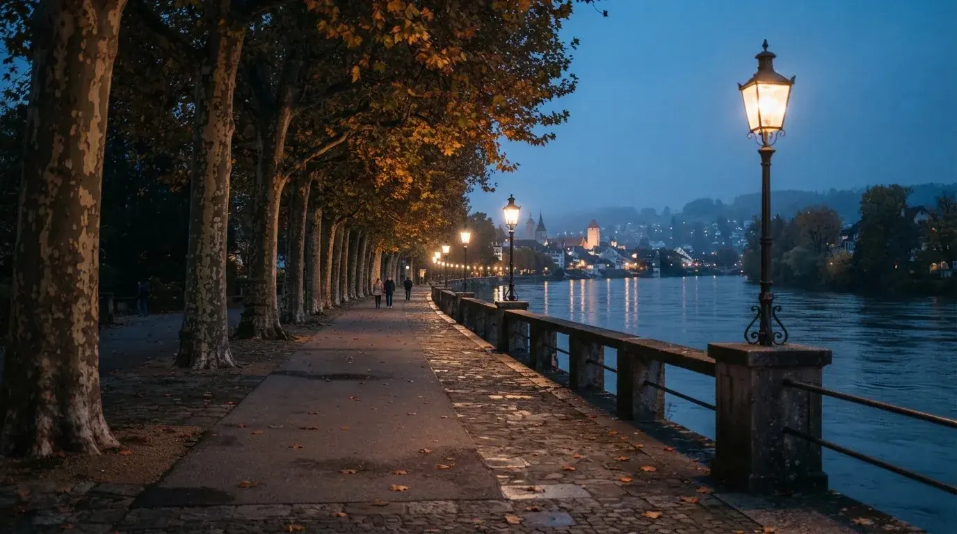 Eine Rheinpromenade mit hohen Bäumen am Abend, das Wasser spiegelt das Licht der ersten Laternen.