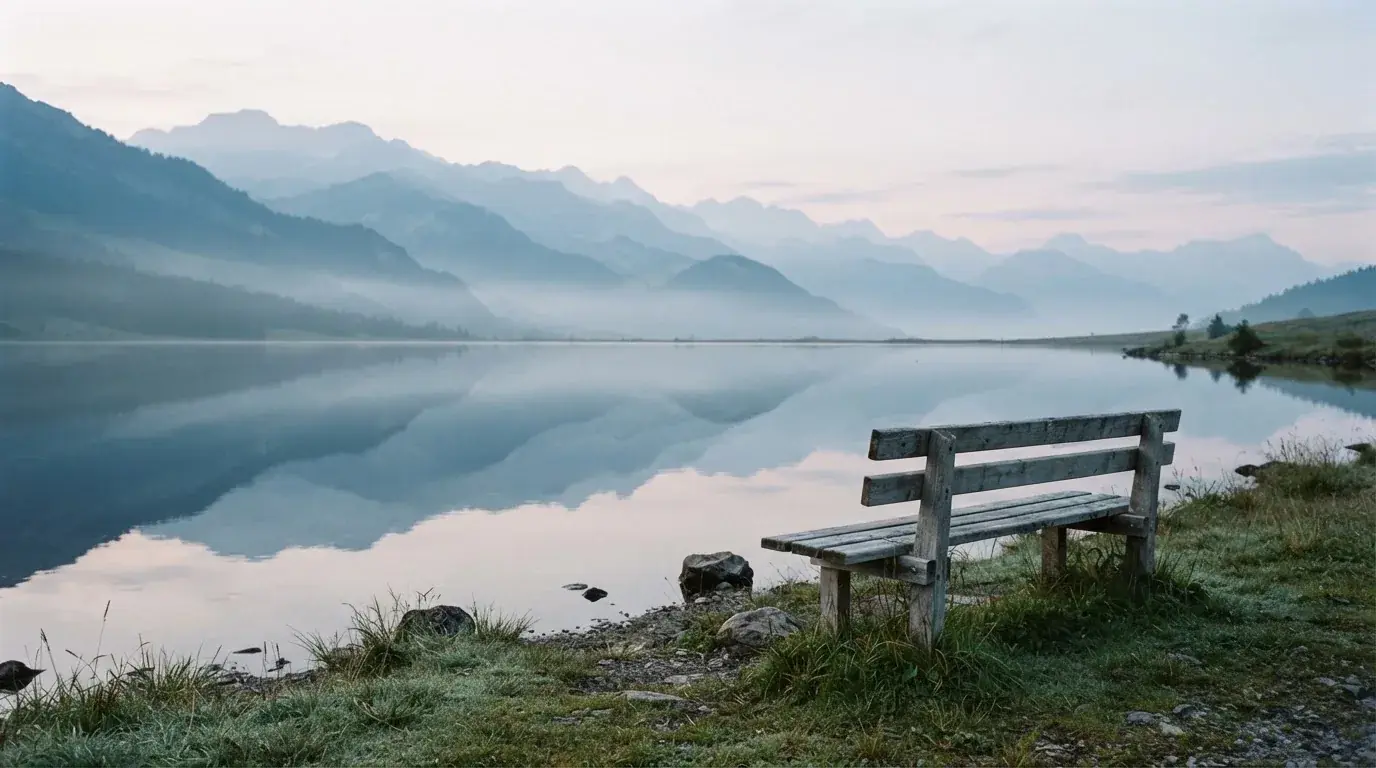 Eine Holzbank an einem Seeufer mit Blick auf eine Bergkette im Morgendunst, das Wasser ist spiegelglatt.
