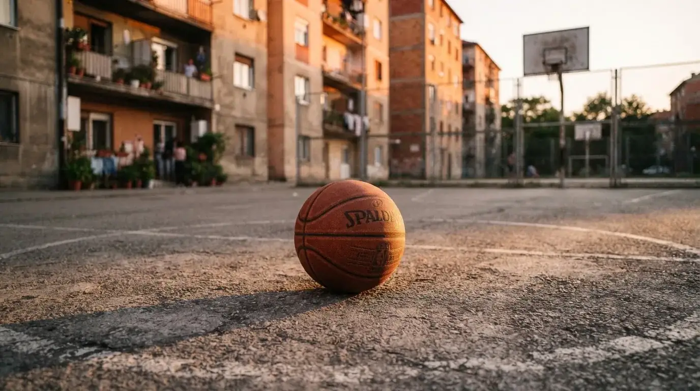 Ein Basketball liegt auf einem Asphaltplatz in einem Hinterhof zwischen Wohnblöcken, das Abendlicht färbt die Wände warm.
