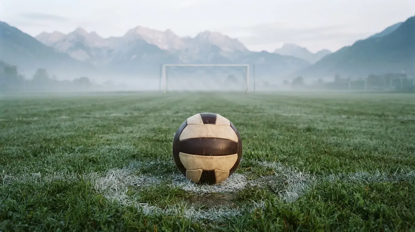Ein einzelner Fussball auf dem Mittelpunkt eines leeren Rasens im Morgennebel, hinter dem Tor ragen Berge auf