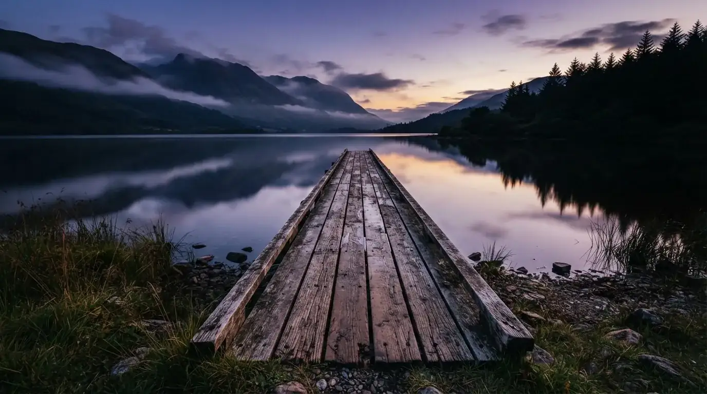 Ein Bergsee in der Dämmerung mit einem leeren Holzsteg, die Wasseroberfläche ist ruhig und Nebel zieht über die Hänge.