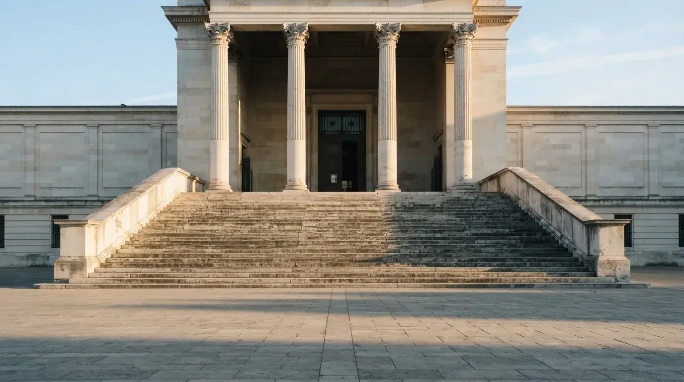 Eine steinerne Treppe führt zu einem Portal mit Säulen im Morgenlicht, der Platz davor ist leer.